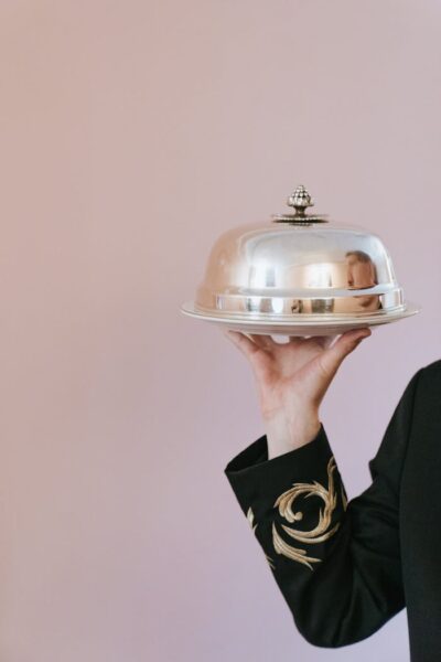 Close-up of a waiter holding a silver serving tray on a light pink background.