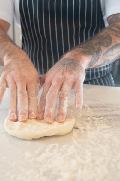 A chef with tattoos kneads dough on a floured surface in a kitchen setting.